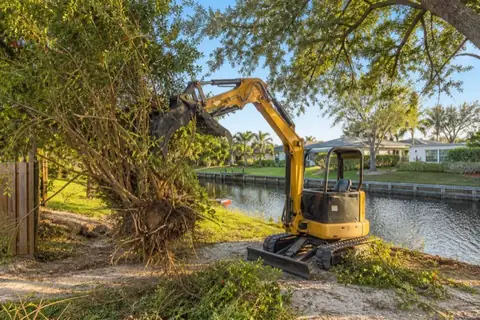 Mini excavator lifting Brazilian pepper stems from a fenced canal-side corner in Central Broward